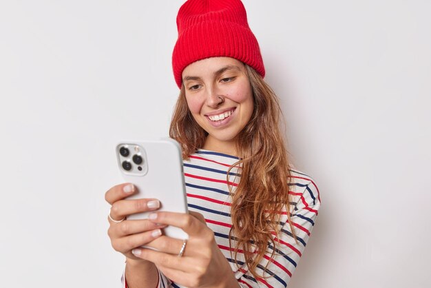 Une adolescente heureuse porte un chapeau rouge et un pull rayé décontracté a un piercing sur le nez tient un téléphone portable fait un appel vidéo communique avec un ami en ligne isolé sur blanc. La technologie