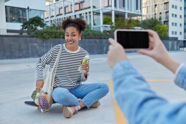 Une adolescente heureuse pose à l'appareil photo d'un smartphone pour faire de la photo est assise les jambes croisées sur une planche à roulettes porte un sac en filet avec des fruits contient une boisson fraîche de désintoxication à base d'agrumes passe du temps libre en milieu urbain