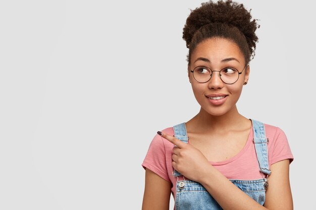 Adolescente joyeuse à l'air agréable avec des cheveux afro nets, pointe avec l'index dans le coin supérieur gauche, montre un espace libre pour le contenu promotionnel, porte un t-shirt décontracté, des lunettes pour une bonne vision