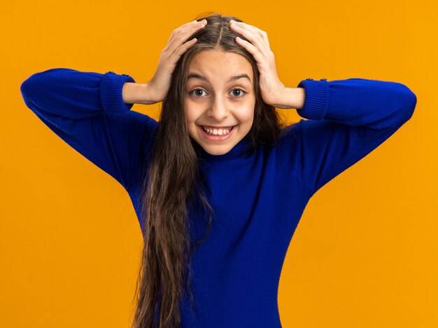 Adolescente joyeuse gardant les mains sur la tête regardant l'avant isolé sur le mur orange