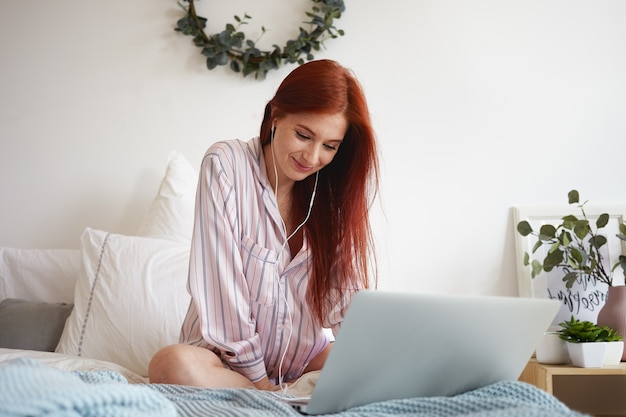 Adolescente mignonne aux cheveux roux naturel paressant dans son lit pendant la journée, assise devant un ordinateur portable ouvert portant un costume de nuit et des écouteurs, profitant des morceaux de musique préférés à l'aide d'un service en ligne gratuit