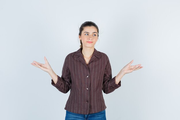 Adolescente montrant un geste impuissant, regardant loin en chemise rayée marron et semblant indécise. vue de face.