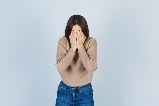 Adolescente en pull, jeans couvrant le visage avec les mains et l'air déprimé, vue de face.