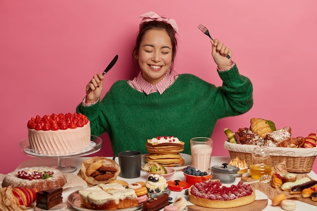 Photo gratuite une adolescente ravie bénéficie d'un événement festif, s'assoit à table avec divers gâteaux gastronomiques, des boissons et des biscuits tient un couteau et une fourchette obtient des émotions agréables après avoir somnolé de sucre.