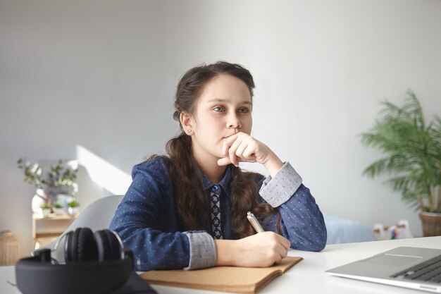 Adolescente réfléchie en chemise décontractée assis au bureau à la maison avec un ordinateur portable et un casque, faire ses devoirs, rédiger un essai en cahier