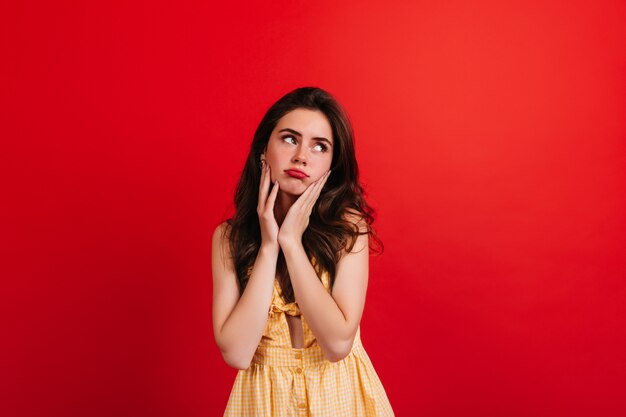 Adolescente en robe d'été jaune posant de mauvaise humeur contre le mur rouge. Closeup portrait de femme brune aux lèvres rouges.