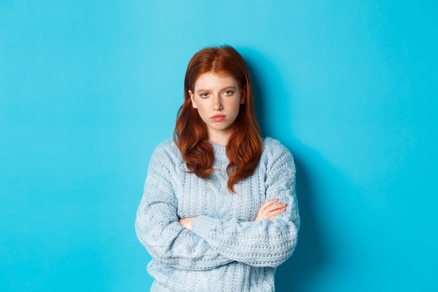 Une adolescente rousse agacée et dérangée croise les bras sur la poitrine, regardant quelque chose de boiteux et d'ennuyeux, debout sur fond bleu