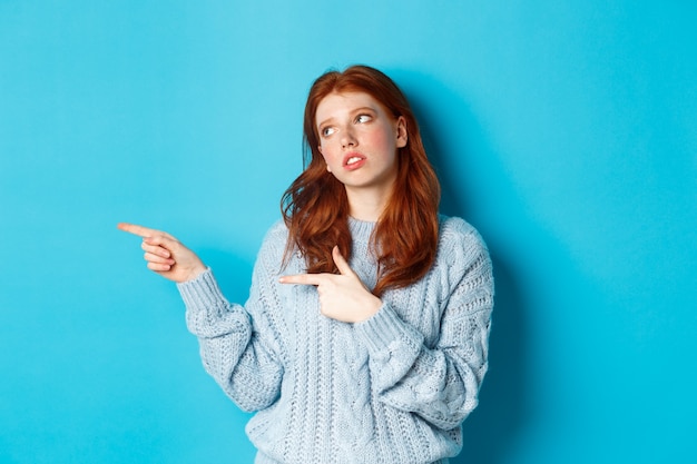 Une adolescente rousse agacée roule les yeux, pointant du doigt quelque chose d'ennuyeux ou de boiteux, debout irritée sur fond bleu.