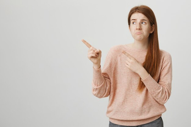 Adolescente rousse sombre bouleversée pointant les doigts dans le coin supérieur gauche et faisant la moue