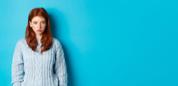 Adolescente rousse triste et sombre regardant la caméra mal à l'aise se sentant mal debout contre le dos bleu