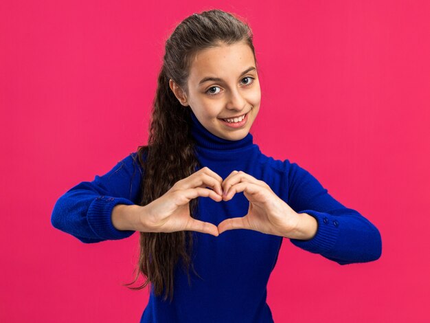 Adolescente souriante faisant un signe de coeur regardant devant faisant un signe de coeur isolé sur un mur rose