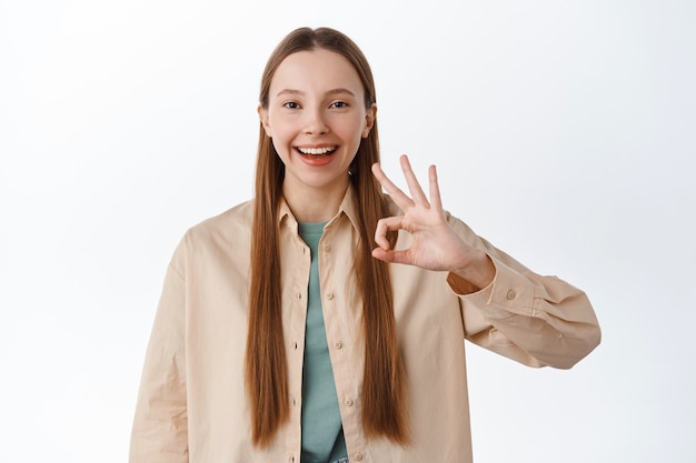 Une adolescente souriante et satisfaite avec une coiffure longue naturelle et un maquillage nude, montrant un geste OK, hochant la tête en signe d'approbation, dites oui, aimez et acceptez, louez un excellent choix, mur blanc.