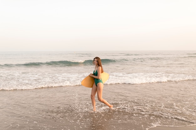 Adolescente souriante tenant la planche de surf, marchant près de la côte à la plage