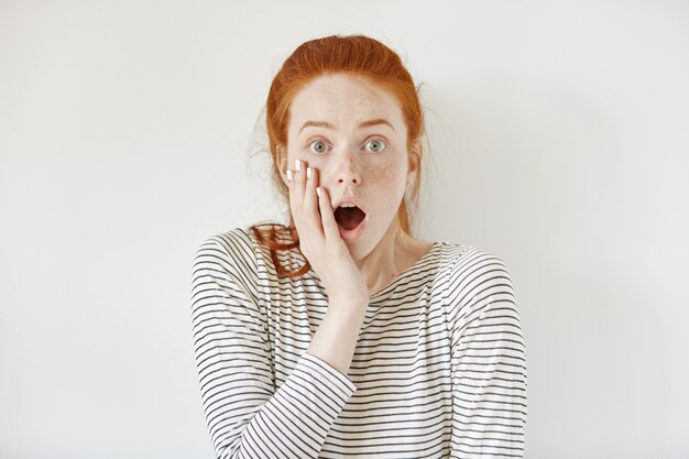 Adolescente surprise, étonnée, portant une chemise de marin touchant sa joue et gardant la bouche grande ouverte, choquée par des nouvelles inattendues. Oublieuse jeune femme à la peur et peur