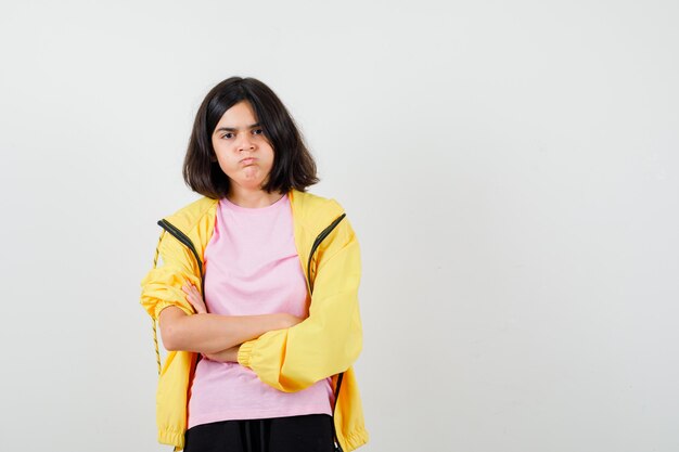 Adolescente en survêtement jaune, t-shirt debout avec les bras croisés, gonflant les joues et l'air insatisfait, vue de face.