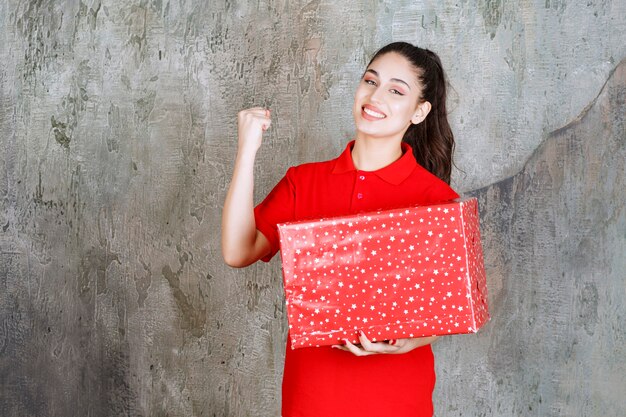 Adolescente tenant une boîte-cadeau rouge avec des points blancs dessus et montrant son poing