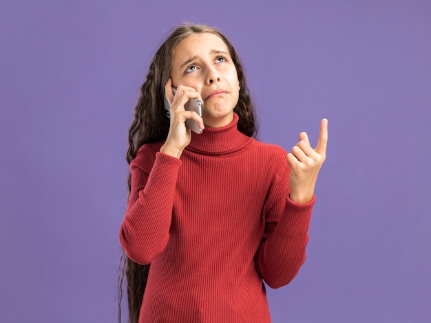 Adolescente triste parlant au téléphone regardant et pointant vers le haut isolé sur un mur violet avec espace de copie