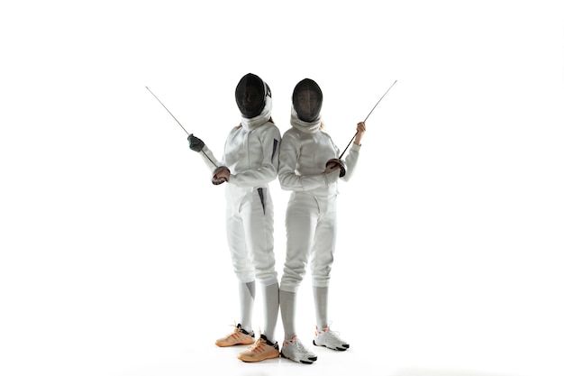 Les adolescentes en costumes d'escrime avec des épées dans les mains isolés sur fond de studio blanc. Formation de jeunes modèles féminins, posant confiant. Copyspace. Sport, jeunesse, mode de vie sain, mouvement, action.