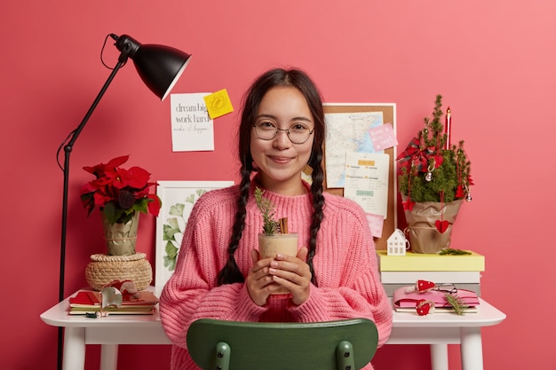 Adorable Adolescente Brune Vêtue D'un Pull D'hiver, Tient Le Lait De Poule à La Cannelle, Porte Des Lunettes Rondes, S'assoit Sur Une Chaise Près Du Lieu De Travail, La Couleur Rose Prévaut.