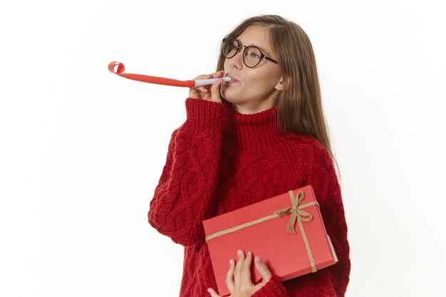 Adorable charmante jeune femme portant des speactales et un pull soufflant la corne de fête tout en s'amusant, célébrant son anniversaire, tenant un cadeau dans une boîte rouge avec ruban. Concept de célébration et de vacances