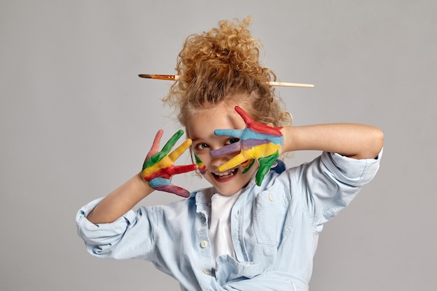 Photo gratuite adorable écolière ayant une brosse dans ses cheveux blonds bouclés chics, vêtue d'une chemise bleue et d'un t-shirt blanc. elle regarde à travers ses mains peintes et sourit, sur un fond gris.