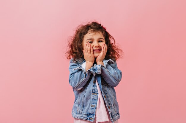 Adorable enfant mignon en veste en jean touchant le visage. Sourire fille préadolescente posant sur fond rose.