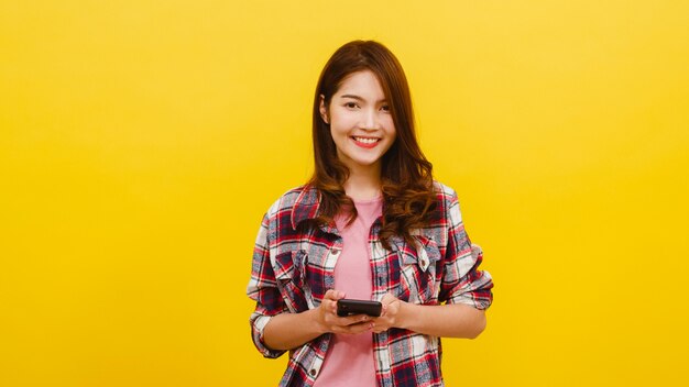 Adorable femme asiatique souriante utilisant un téléphone avec une expression positive, sourit largement, vêtue de vêtements décontractés et regardant la caméra sur le mur jaune. Heureuse adorable femme heureuse se réjouit du succès.