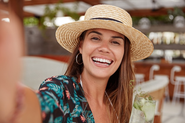 Adorable femme au chapeau de paille, porte un chemisier, fait un selfie.