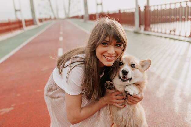 Adorable femme aux cheveux bruns souriant avec son chien en marchant sur la ville du matin