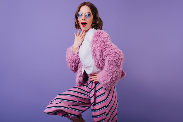 Adorable femme blanche à lunettes de soleil posant avec une expression de visage surpris sur le mur violet. Plan intérieur d'une fille glamour étonnée portant un pantalon rose et une veste de fourrure.