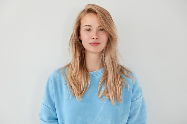 Adorable femme blonde avec une expression sérieuse, vêtue d'un pull bleu, a une peau saine et propre, isolée sur un mur blanc. Jolie femme démontre sa beauté naturelle