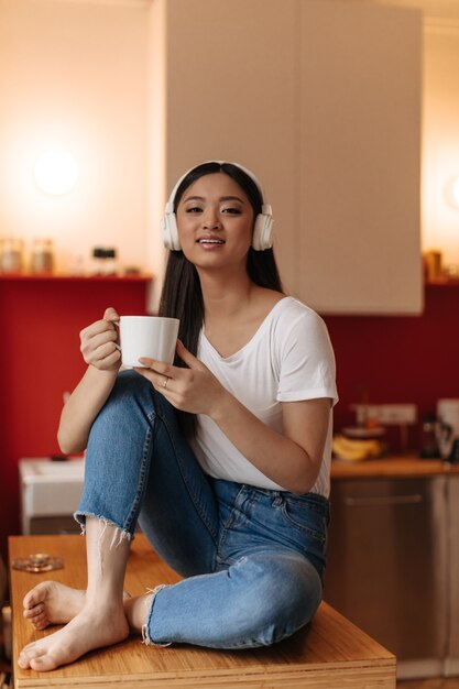 Adorable femme brune dans les écouteurs assis dans la cuisine avec une tasse de thé