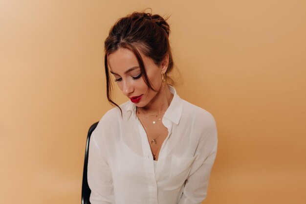 Adorable femme charmante avec faisceau sur la tête avec des lèvres rouges portant une chemise blanche regarde vers le bas et souriant sur fond beige isolé