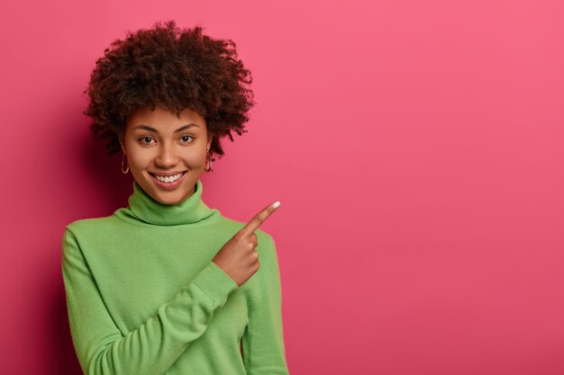 Photo gratuite adorable femme avec une coiffure frisée, une peau saine et des dents blanches, des points sur un endroit vide, montre de l'espace pour votre contenu publicitaire