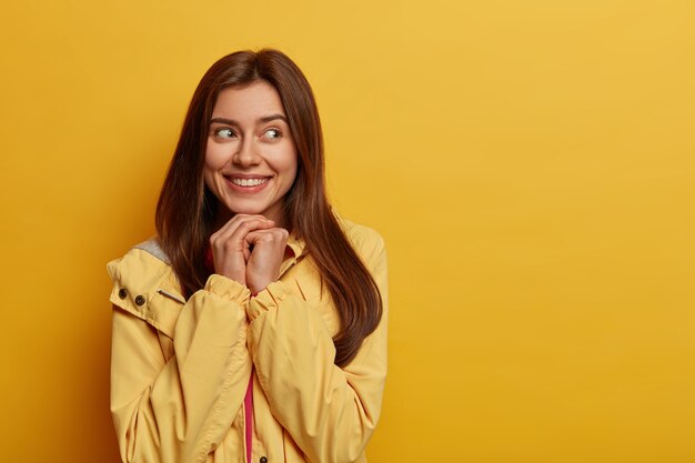 Adorable femme joyeuse a un sourire doux, se souvient d'une situation réconfortante dans la vie, garde les mains sous le menton, a un regard intéressant et intrigué de côté