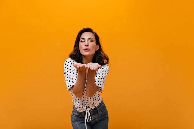 Adorable femme souffle baiser sur fond orange. Dame aux cheveux noirs en chemisier à pois blanc et jean bleu pose pour la caméra.