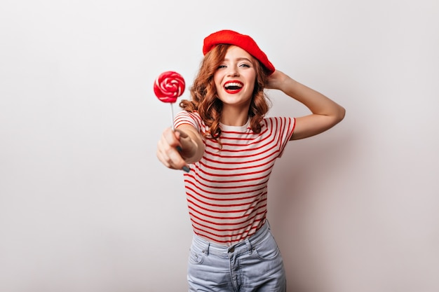 Adorable fille au béret rouge, manger des bonbons. incroyable dame française aux cheveux roux posant avec sucette.