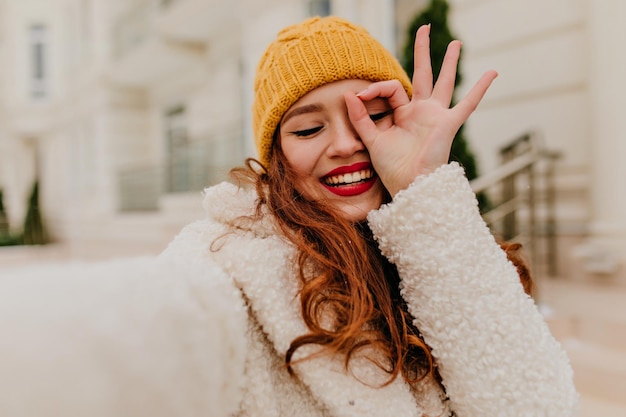 Adorable fille au gingembre faisant du selfie en hiver Femme riante aux cheveux longs en chapeau jaune s'amusant le week-end de décembre