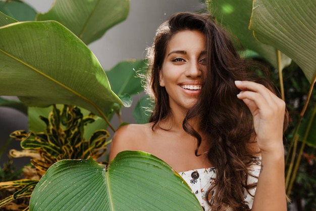 Adorable fille aux cheveux bouclés rit sur fond de grandes feuilles vertes Dame aux yeux verts en haut blanc avec imprimé floral posant