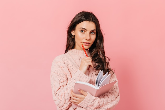 Adorable fille aux yeux bleus posant pensivement sur fond rose. Dame aux cheveux bouclés tenant un crayon rouge et un journal.