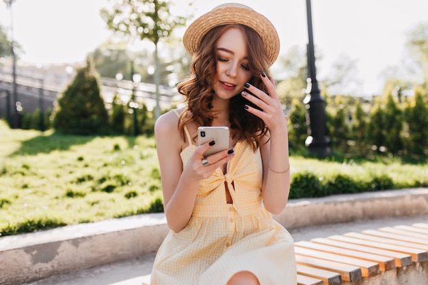 Adorable fille blanche avec manucure noire se détendre dans le magnifique parc d'été. Photo extérieure d'un modèle aux cheveux roux gracieux utilisant son smartphone pendant la séance photo.