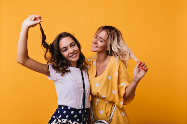 Adorable fille brune touchant ses cheveux et riant. Blithesome soeurs caucasiennes drôles de danse sur lumineux.