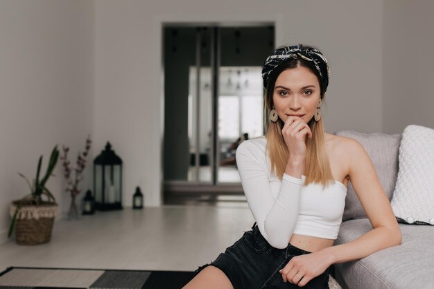 Adorable fille élégante aux cheveux blonds avec accessoire dans la tête portant un haut blanc et un short touche ses lèvres et pose à la caméra tout en se reposant à horm