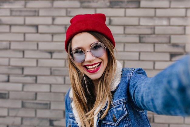 Adorable fille à lunettes bleues faisant selfie avec une expression de visage inspirée. Photo de la belle jeune femme au chapeau prenant une photo sur le mur de briques.