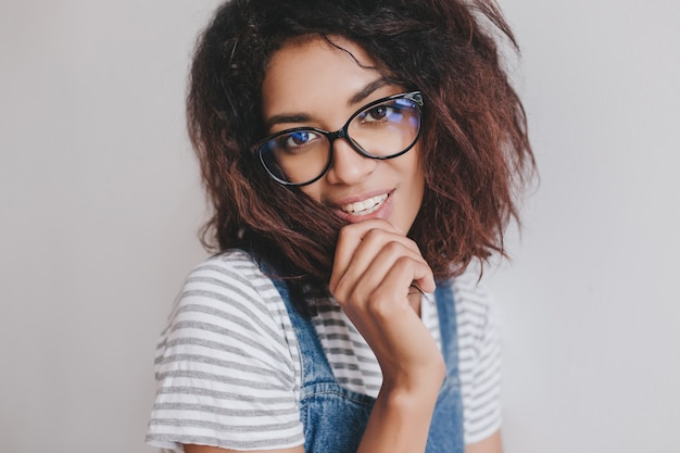 Adorable fille noire avec une coiffure à la mode posant de manière ludique sur un mur lumineux, regardant à travers des lunettes élégantes