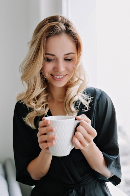 Adorable fille souriante détendue sourit et tient une tasse de café du matin Photo intérieure d'une femme incroyable assise à la maison et se reposant près de la fenêtre