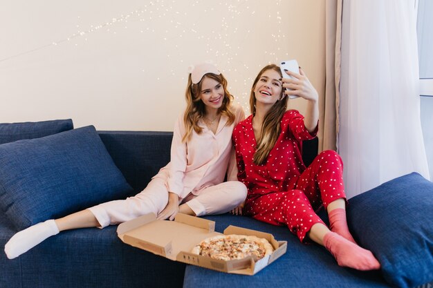 Adorable jeune femme bouclée en chaussettes blanches souriant pendant que son amie fait selfie à la maison. Photo à l'intérieur de deux sœurs ravies en train de manger une pizza le week-end.
