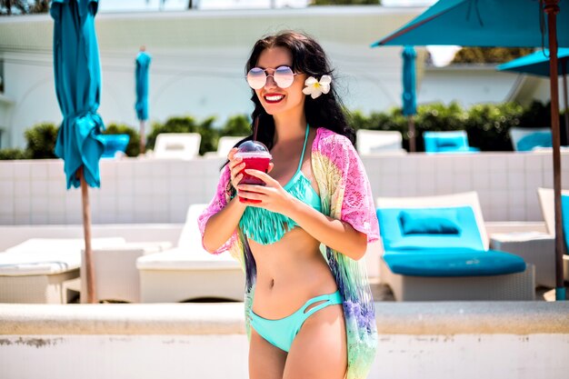 Adorable jeune femme brune sexy posant à la station balnéaire de l'île tropicale, profitez de ses vacances à l'hôtel, journée chaude d'été, boire de la limonade douce et souriant, bikini bleu et accessoires, vêtements de plage à la mode