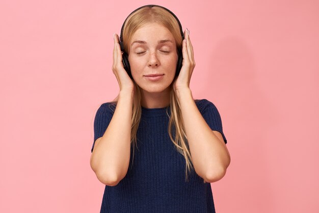 Adorable jeune femme de race blanche avec des taches de rousseur gardant les yeux fermés en profitant du nouvel album de son artiste de musique préféré à l'aide d'écouteurs sans fil