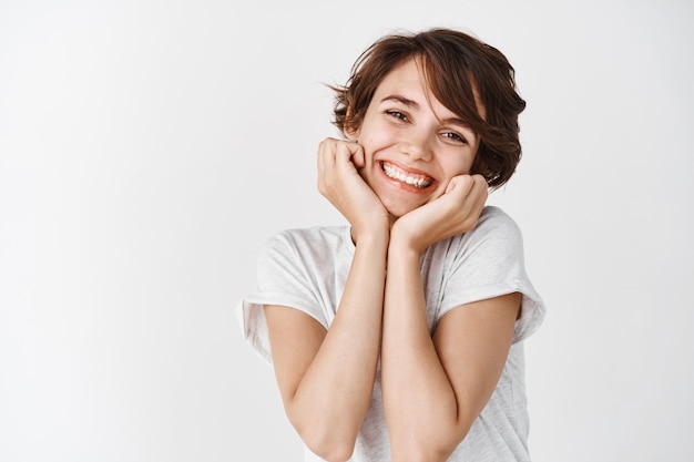 Adorable jeune femme souriante et l'air idiote, montrant une peau éclatante naturelle sans maquillage, debout contre un mur blanc